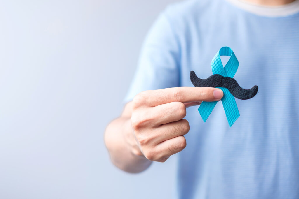 A person wearing a light blue shirt holds a black mustache cutout and a teal ribbon, symbolizing prostate cancer awareness.