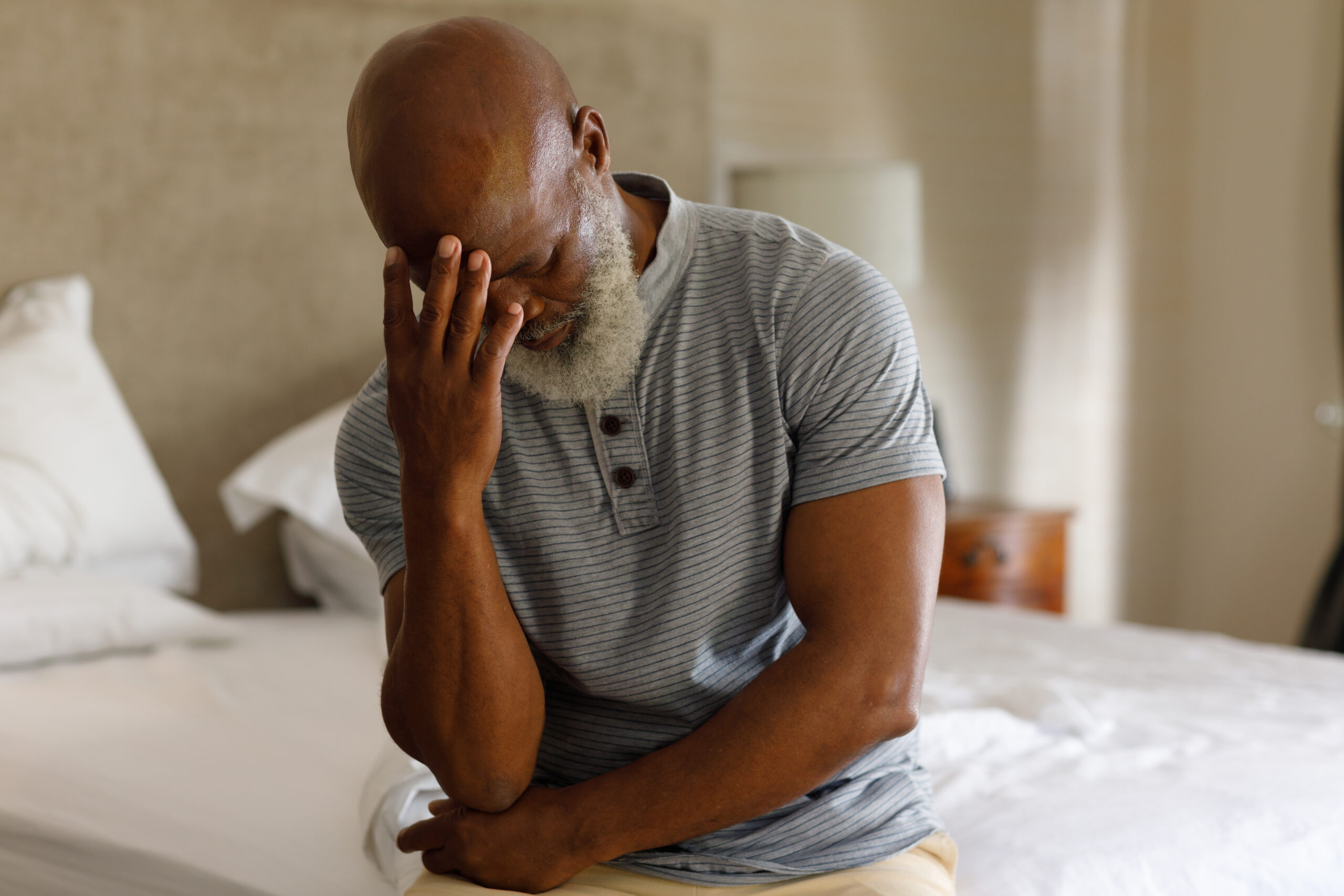 An elderly man with a beard sits at the edge of a bed in his FL home, resting his head in his hand. He appears thoughtful or stressed, perhaps considering a visit to the Men’s Health Clinic in Orlando for some peace of mind.